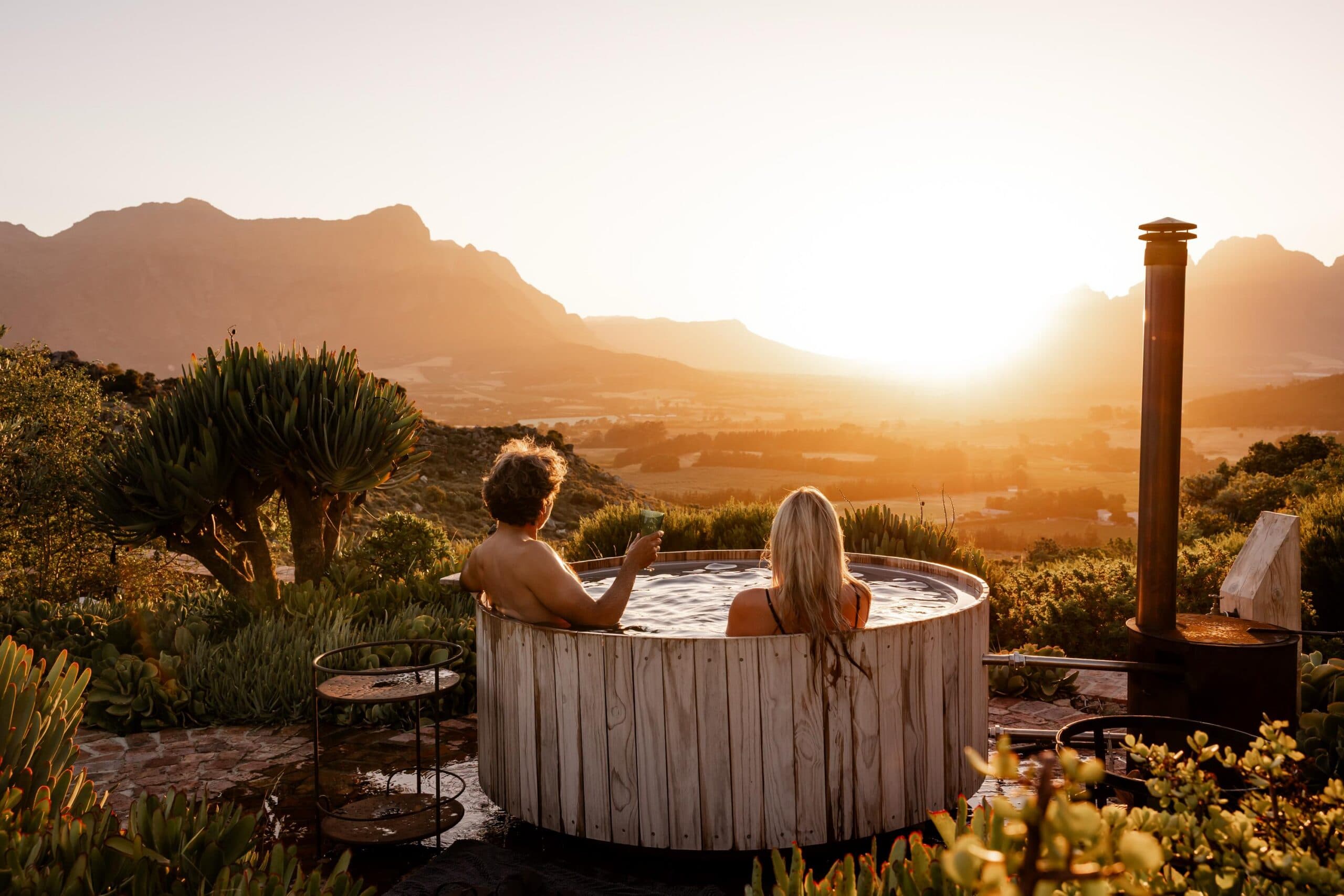 Two people unwind in a round wooden hot tub on a hillside at sunset, overlooking a wide valley framed by mountains. Surrounded by lush shrubs and succulents, one person holds a drink while both face the glowing horizon, with a wood-fired heater beside the tub adding to the tranquil outdoor setting.
